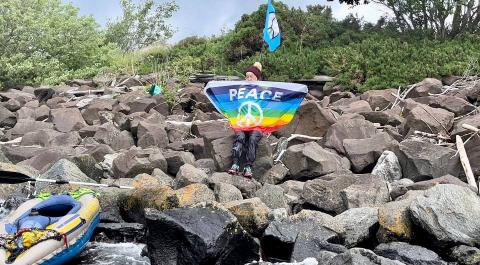 Woman peace activist Ginnie Herbert holds a rainblow flag with a CND symbol on it and the word 'Peace', standing on rocks which are part of the ministry of defence's Coulport nuclear warhead storage facility. Nearby is the inflatable canoe that she used to paddle in and land in the base.
